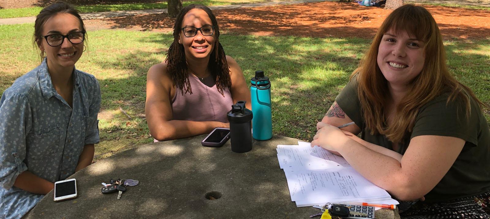 三个微笑, 女, Caucasian students sitting at a round, grey stone table: the first has brown hair with blond highlights worn down and is wearing a black shirt and a black skirt with multi-colored polka dots and is pictured with a stack of paper under her crossed arms on the table with a ring of keys including yellow and white cards and a black cell phone; the second has brown hair worn down and black rimmed glasses with a lavendar pink shirt, 她和一个高个子的合照, 蓝绿色和黑色的水瓶, a shorter solid black water bottle, and her cell phone with a lavendar back and white edges; 和 third has on a blue, collared button up shirt with white polka dots and tan pants, she is pictured with her car keys and a cell phone with white, horizontal bars on the upper and lower portion of the face.